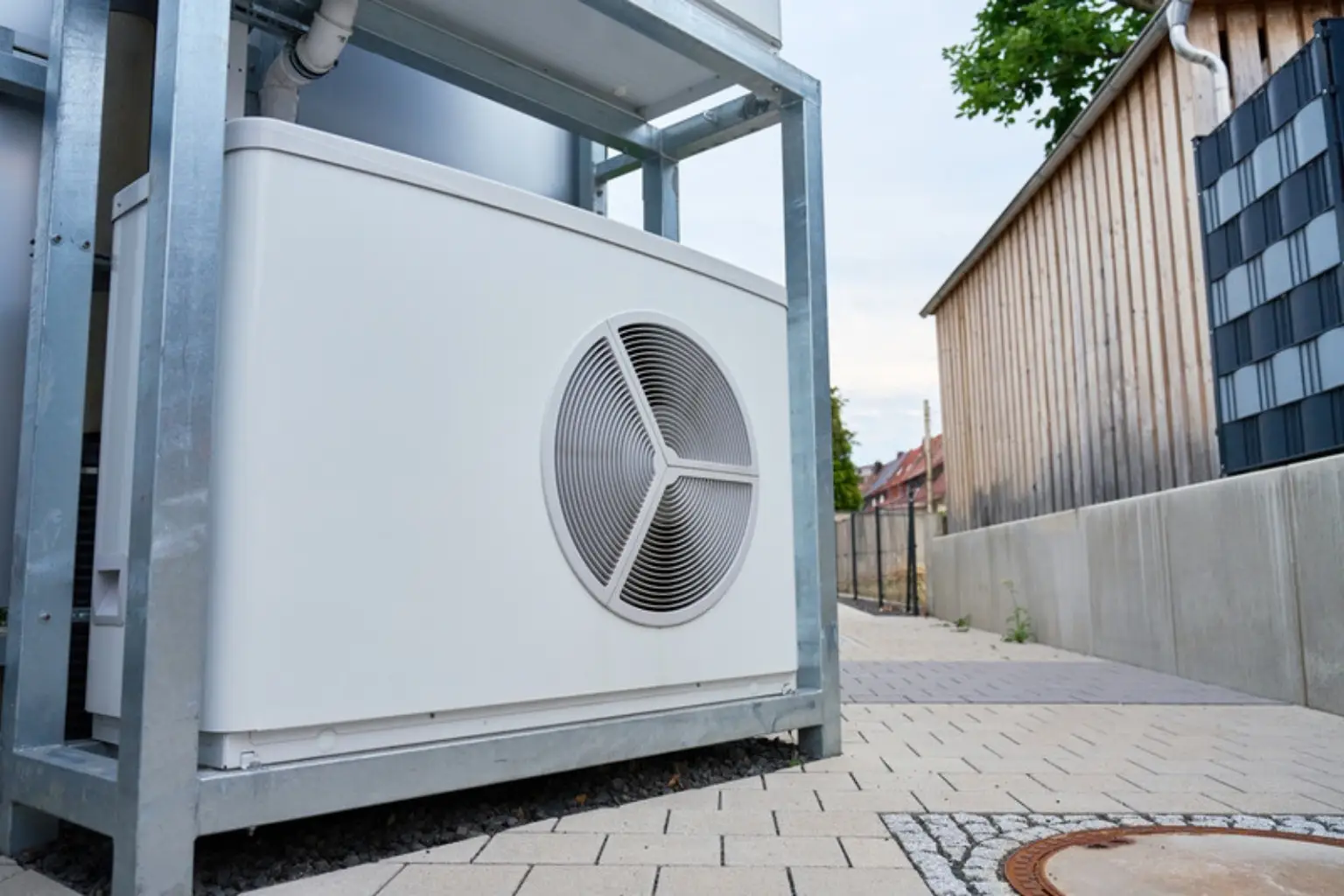 Hvid udedel til varmepumpe på et metalstativ ved en flisebelagt sti